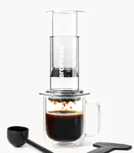 AeroPress Clear coffee maker pressing coffee into a glass mug, with its scoop and stirrer