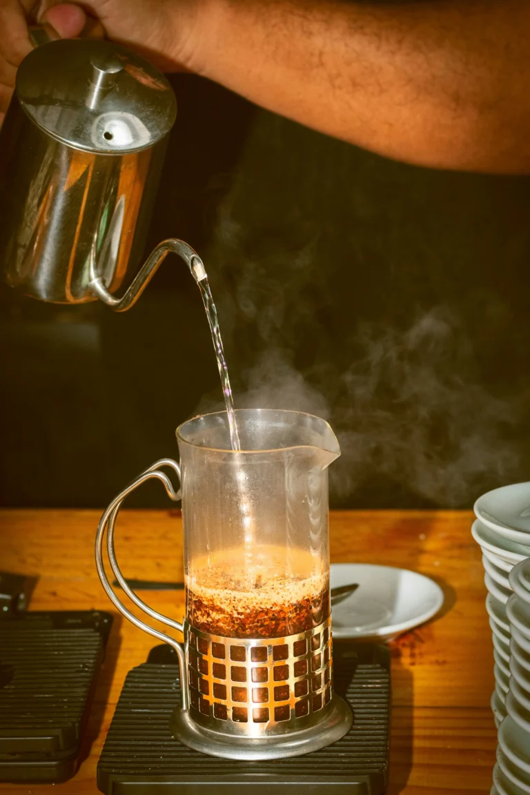Hot water poured from a gooseneck kettle into a glass French press of coffee sitting on scales